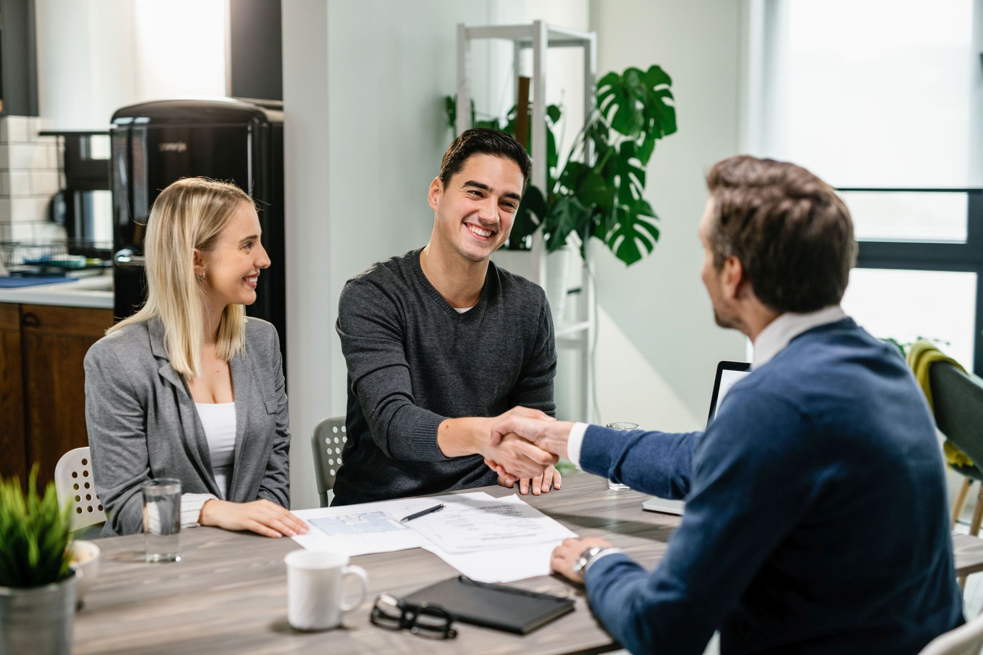 Young happy couple making an agreement with their financial advisor at home. Men are shaking hands. 