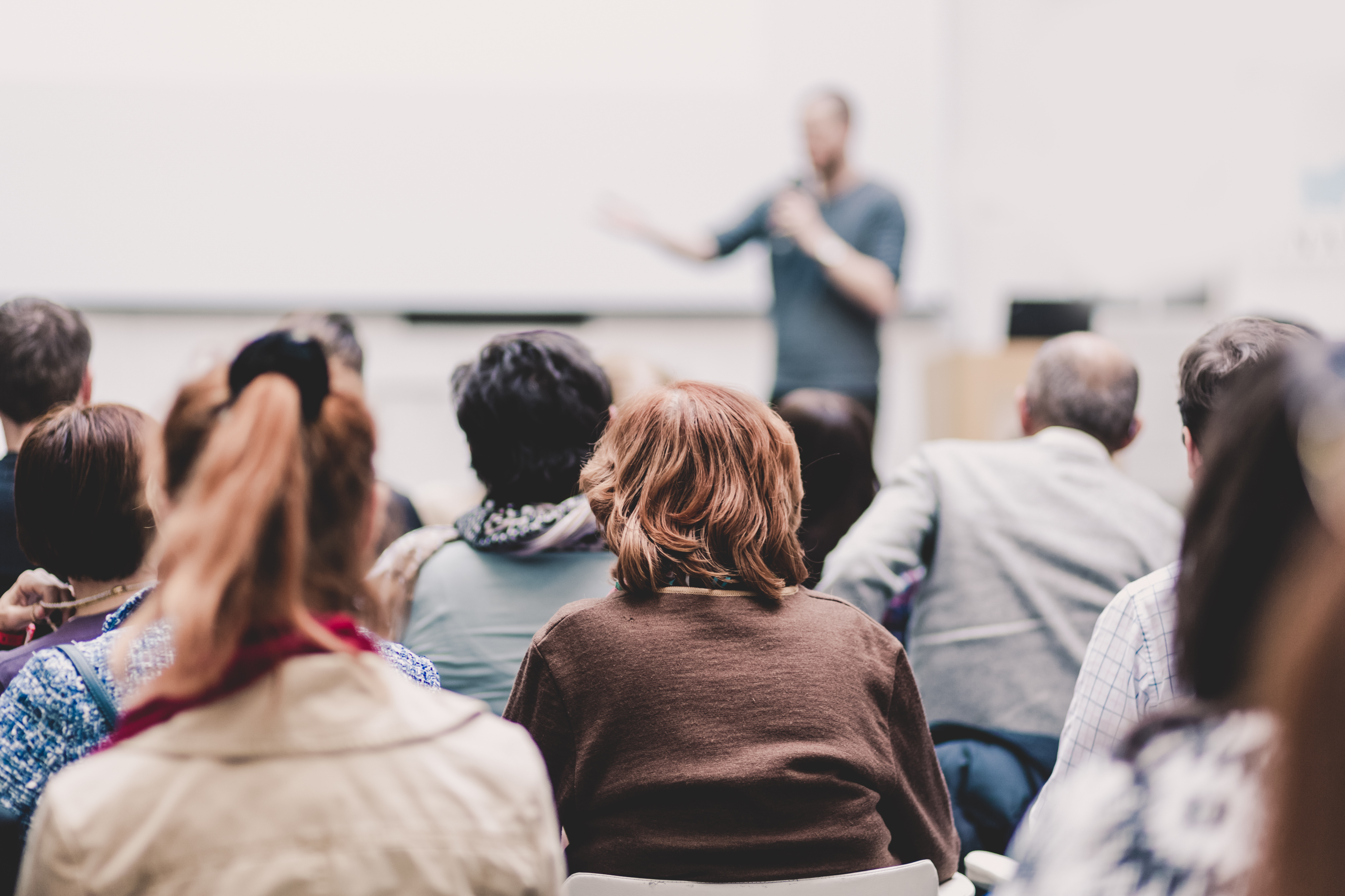 Male speaker giving presentation in lecture hall at university workshop. Audience in conference hall. Rear view of unrecognized participant in audience. Scientific conference event.